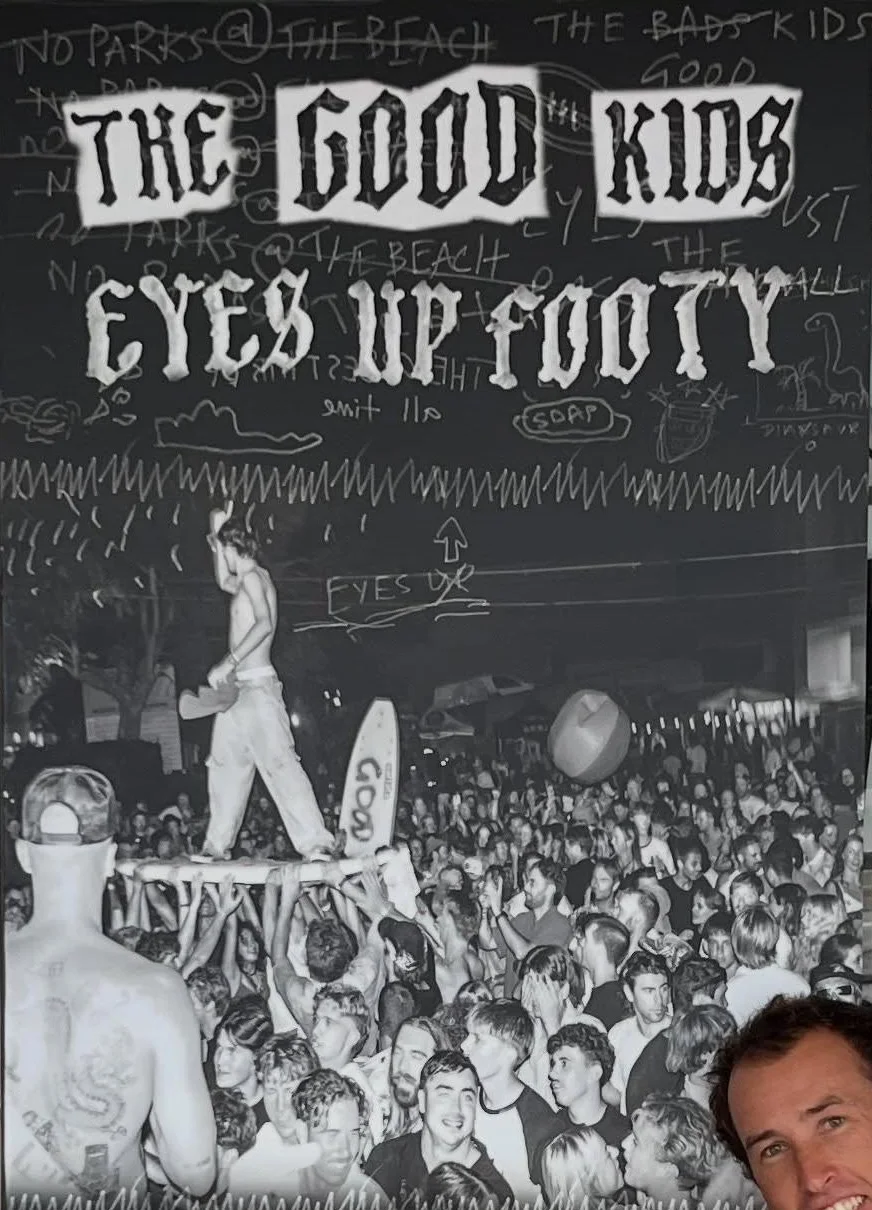 Eyes Up Footy poster: a bloke stands on a surfboard held up by a packed crowd at night, hand-drawn lettering above