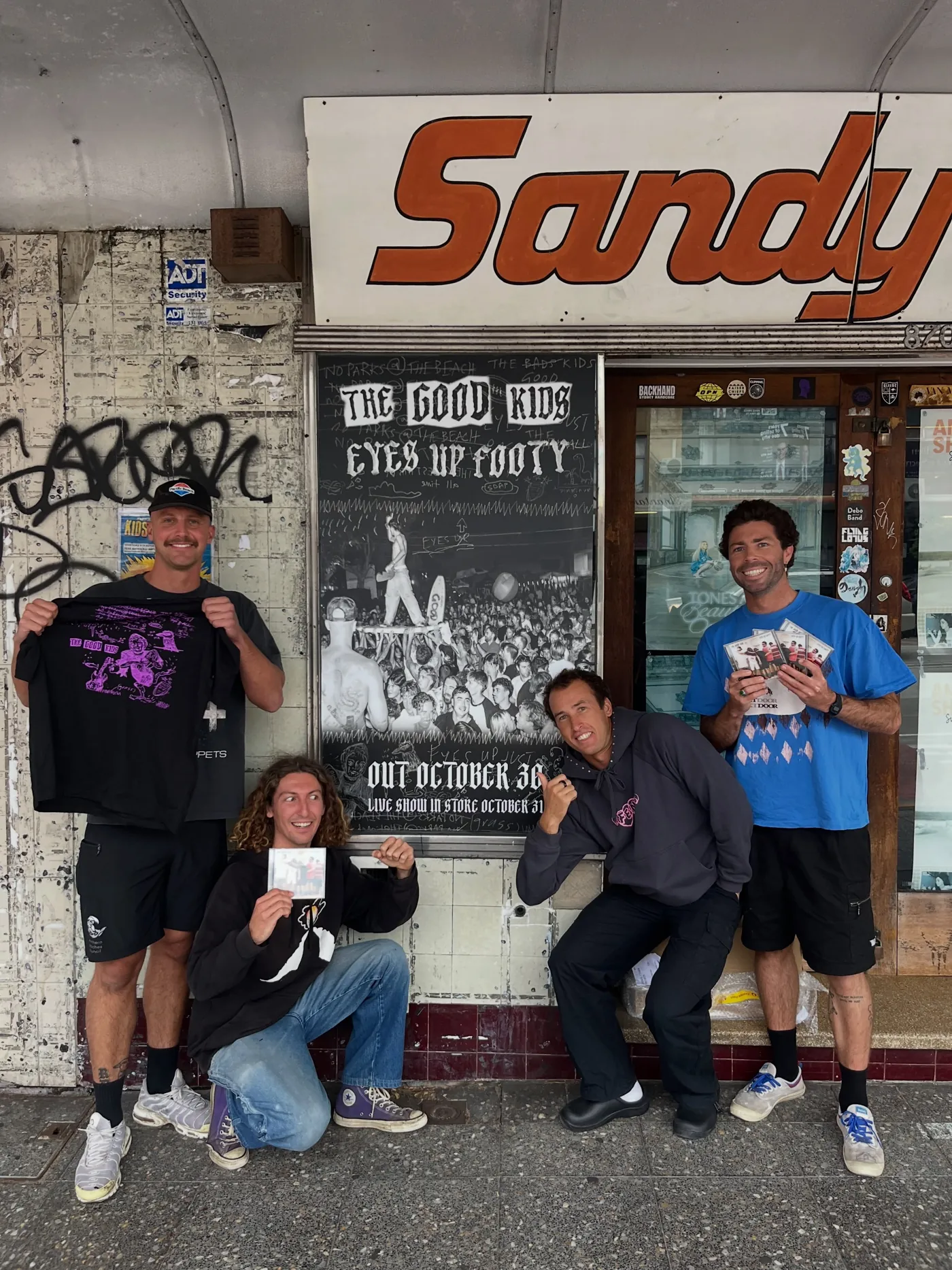 The band outside Sandy's Music in Dee Why holding up the new shirt and CDs beside the Eyes Up Footy poster