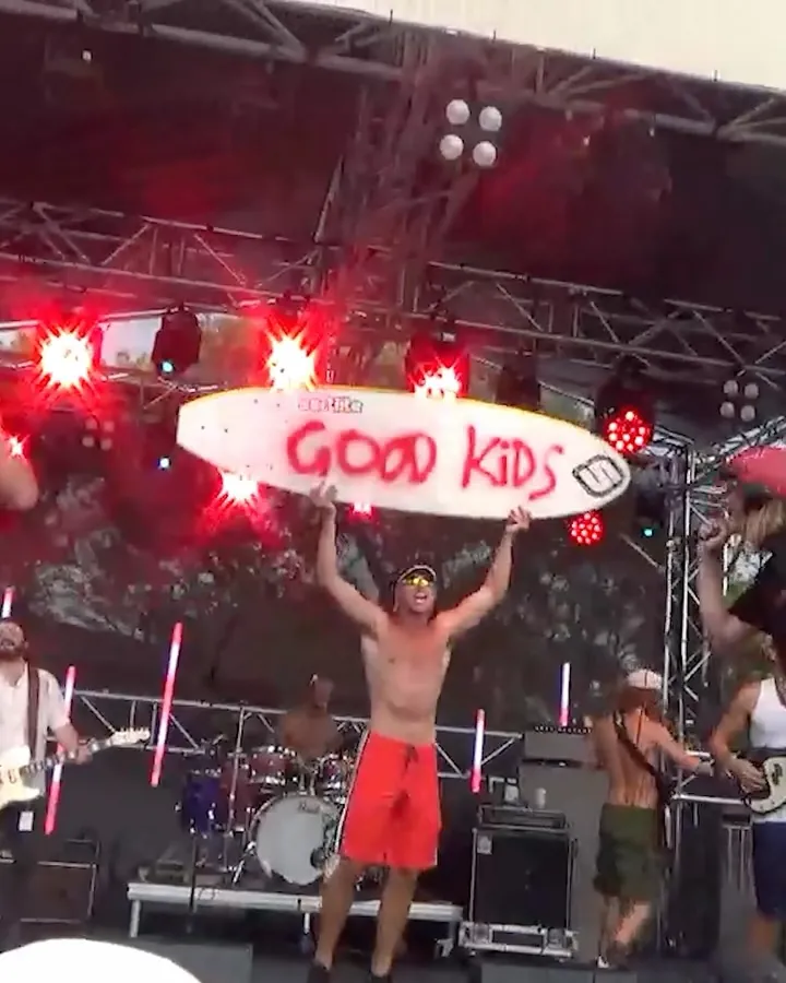 The band on a festival stage holding up a surfboard painted Good Kids